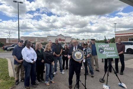 Senator Chuck Schumer speaks outside a Tops grocery store, holding a Senate seal, with a "Lower Prices for Families" sign.