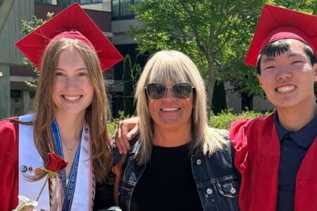 A smiling woman in sunglasses poses with two graduates in red caps and gowns outdoors on a sunny day.
