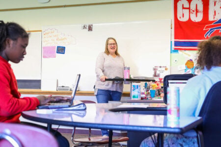 Smiling teacher stands as two students work on laptops in a classroom with a whiteboard and Buffalo Bills flag.