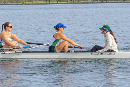 Two William Smith College rowers hold oars with their coxswain in a shell on the water.
