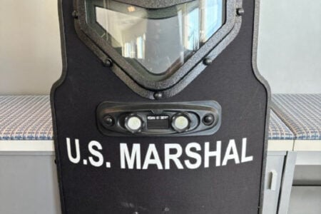 Black ballistic shield with U.S. MARSHAL lettering, a viewport, and lights, standing by an indoor striped bench.