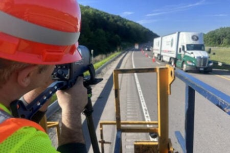 Worker in orange hard hat and safety vest aims a laser speed device at traffic on a highway.
