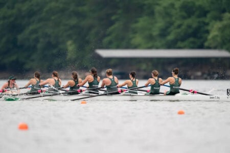 Nine women in a white "James C. Joy" rowing shell, wearing green Herons tops, rowing on a lake.