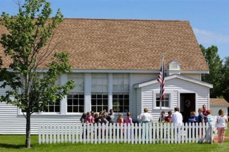 An adult leads a group of children outside a white historic schoolhouse with a picket fence and American flag.