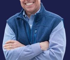 Smiling man with short gray hair, wearing a blue shirt and dark blue vest, standing with arms crossed against a solid dark blue background.