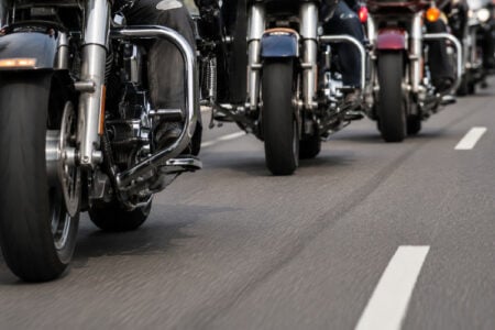 Line of motorcycles with riders traveling on a paved road with white lane markings.