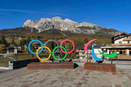 Colorful Olympic rings and Paralympic symbols stand on a plaza, with the Dolomites and a town in the background.