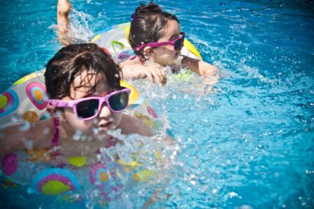 Two girls wearing sunglasses and swim rings splash in a sunny blue pool.