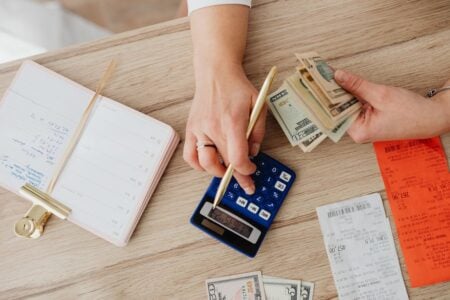 A person's hands calculating on a calculator, counting cash, and handling receipts on a wooden desk.