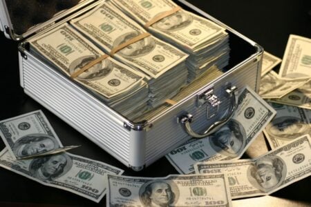 An open silver briefcase overflows with stacks of $100 bills, spilling onto a dark surface.