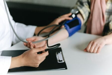 Doctor in a white coat measuring a patient's blood pressure with a cuff and pump at a medical office desk.