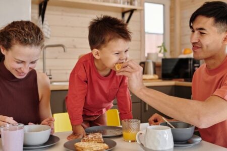 Parents and their young son eat breakfast at a kitchen table, with the father feeding the boy a waffle.