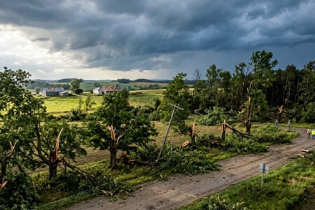 Utility workers assess rural storm damage: fallen trees, broken power lines, and damaged farm roofs under dark clouds.