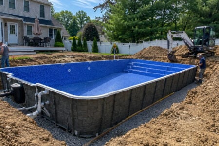 Workers install a blue-lined inground pool in a dirt-dug backyard, with an excavator and a house in the background.