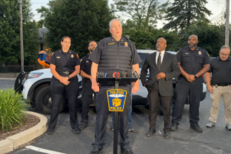Toledo Police Commander speaks at an outdoor press conference, flanked by officers and officials, with a police SUV behind him.