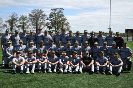 Keuka College baseball team poses for a group photo on their field, wearing grey and green uniforms, with trees and a lake in the background.