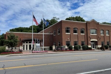 Red brick Victor Town Hall with US & New York flags and a veterans monument, seen from the street under clouds.