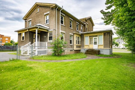 A two-story grey stucco house with yellow trim and a front porch, surrounded by a green lawn and a chain link fence.