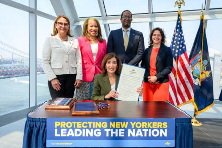 Governor Kathy Hochul holds a signed bill with four officials at a ceremony overlooking a city skyline and water.