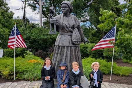 Four children in period attire, including a Union soldier, pose with the Harriet Tubman statue, flanked by US flags in a park.