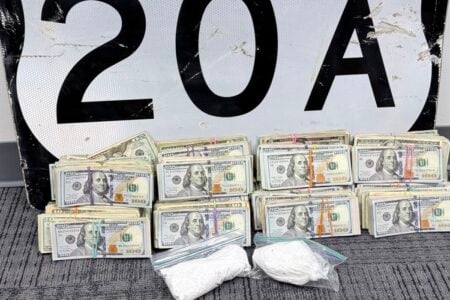 Stacks of US $100 bills and two bags of white powder on a carpet, with a weathered US Route 20A sign behind them.