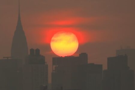 Fiery orange sun obscured by haze over a NYC skyline, silhouetting buildings including the Chrysler Building.