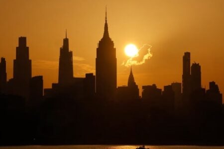 NYC skyline at sunset with the Empire State and Chrysler Buildings silhouetted against an orange sky over water.