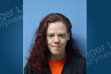 Woman with reddish-brown wavy hair and an orange shirt posing for a mugshot against a light blue background.