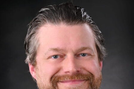 Smiling man with a reddish-brown beard and blue eyes in a dark suit jacket and white shirt, against a dark studio background.