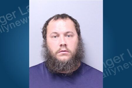 Man with a full dark beard and short, dark hair poses for a mugshot against a light background.