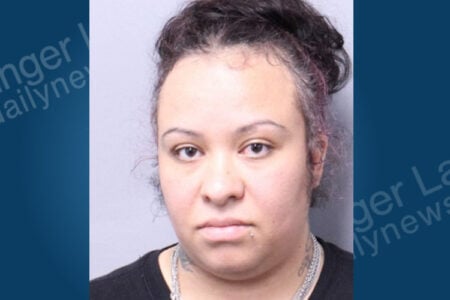 Woman with dark, curly hair, a lip piercing, and neck tattoos poses for a police mugshot.