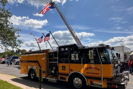 Aurelius Fire Department's orange and black fire truck with an American flag on its extended ladder at an outdoor event.