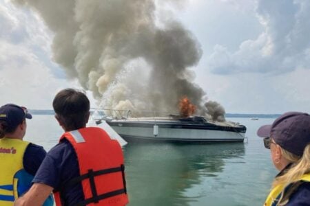 Three people on a boat, one holding a hose, observe a large boat engulfed in flames on a lake.