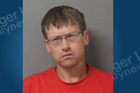 A man with light brown hair, glasses, and a red shirt poses for a mugshot against a plain gray background.