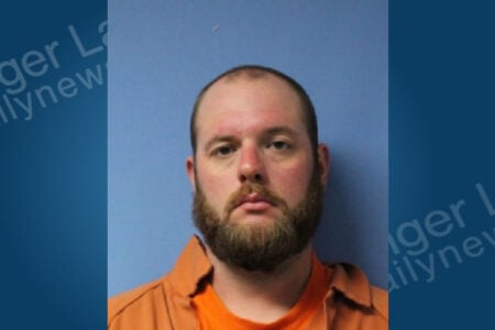Mugshot of a bearded man with a receding hairline wearing an orange shirt against a blue background.