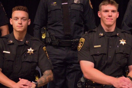 Two Onondaga County Sheriff's deputies in dark uniforms smile while seated indoors.