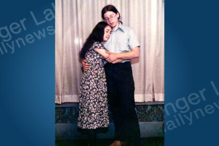A young woman in a floral dress embraces a young man in a light shirt and glasses, standing against a curtain.