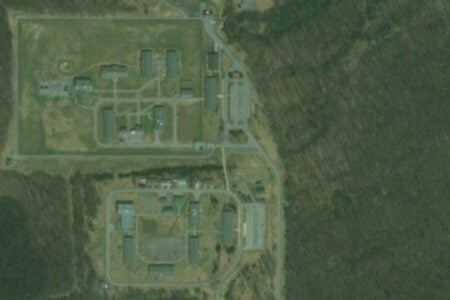 Aerial view shows a large correctional facility with fenced compounds, numerous buildings, and roads, surrounded by dense forest.