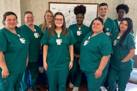 Ten smiling UR Medicine Finger Lakes CNA students from the 2023 class pose indoors in teal scrubs.