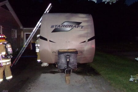Two firefighters stand near a scorched StarCraft travel trailer on a dark driveway by a house at night.