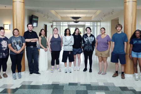 Eleven diverse young adults stand in a line in a bright, modern hallway with columns.