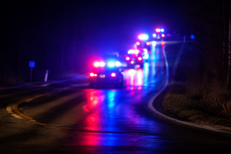 Out-of-focus police cars with flashing red and blue lights on a wet, dark, winding road at night.