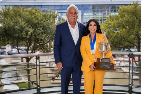 Smiling man and woman on outdoor balcony overlooking water. Woman in yellow suit holds large silver trophy and wears a medal.