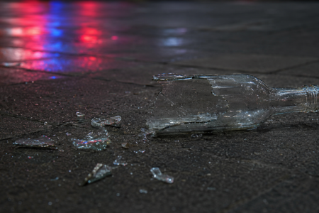 Broken clear glass bottle and shards on dark, wet pavement with colorful light reflections.