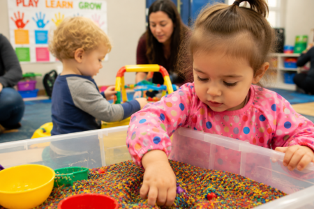 A toddler girl plays with colorful sensory rice in a bin at a preschool, supervised by adults.
