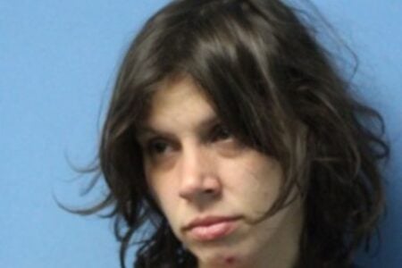 Mugshot of a young woman with dark, messy hair and an orange shirt over a dark uniform, against a blue background.