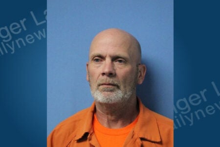 Bald man with a grey beard wearing an orange top, looking straight ahead against a blue background.
