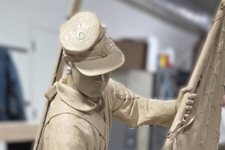 Clay sculpture of a Civil War soldier wearing a cap with "111 G" holding an American flag in a studio.