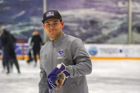 Man wearing a Niagara Hockey cap and jacket smiles on an ice rink, holding a hockey stick and purple glove.