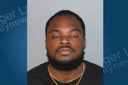 Bearded Black man with diamond earrings and gold chains, wearing a black shirt, in a mugshot against a plain background.